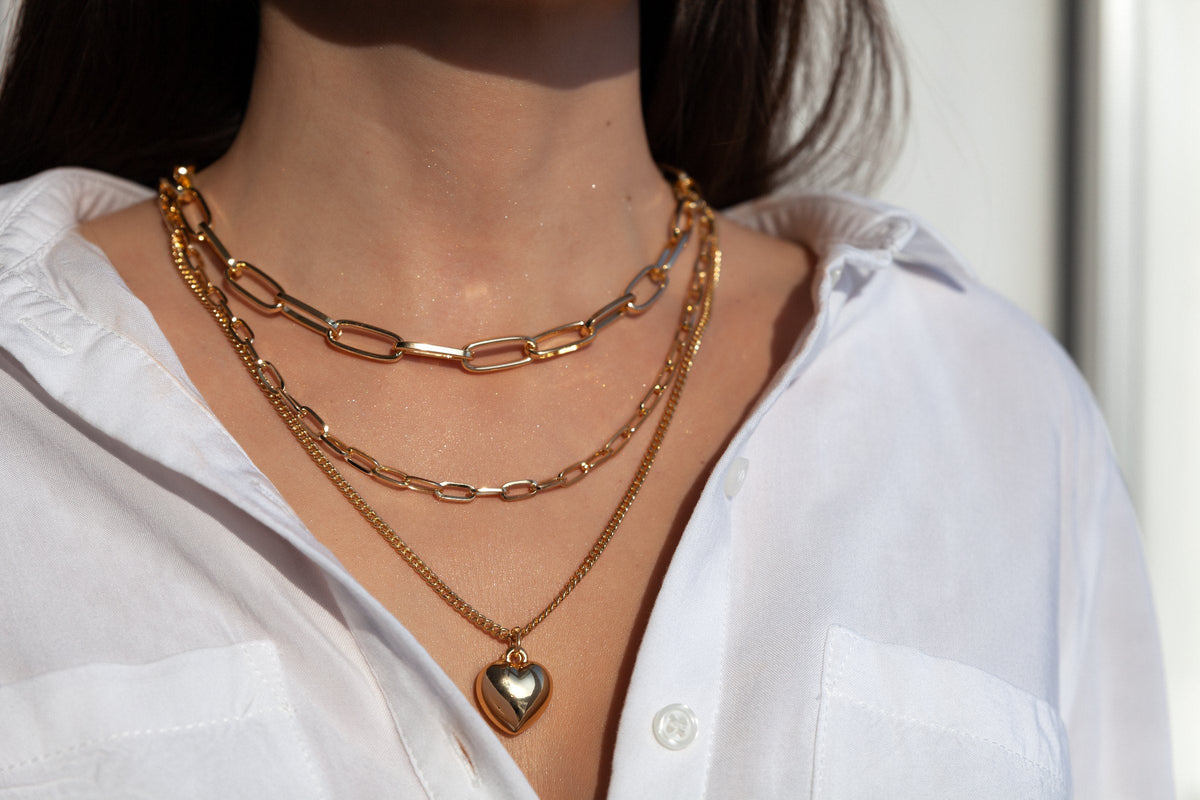 Layered gold chain necklaces ending in a heart pendant, worn over an open white shirt