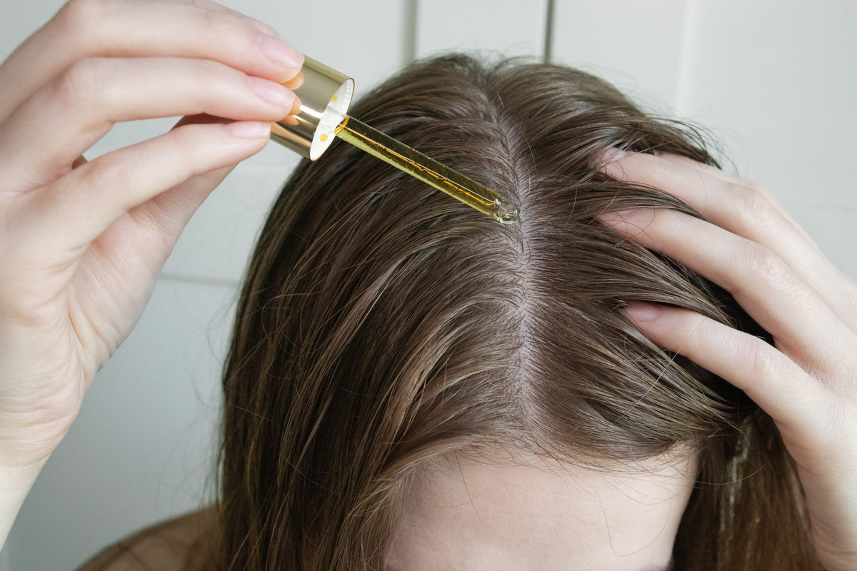 A woman parting her hair and applying a few drops of serum to her scalp with a glass dropper