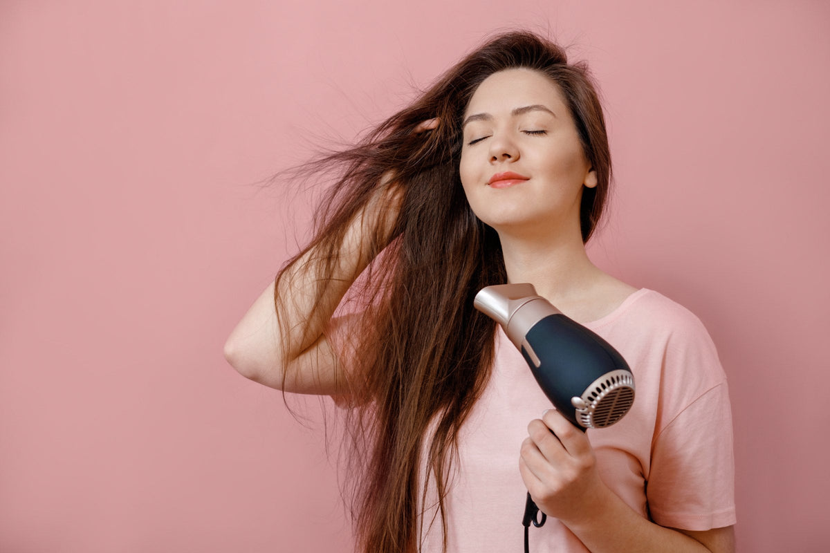 A young woman blow-drying her hair for volume against a pink background