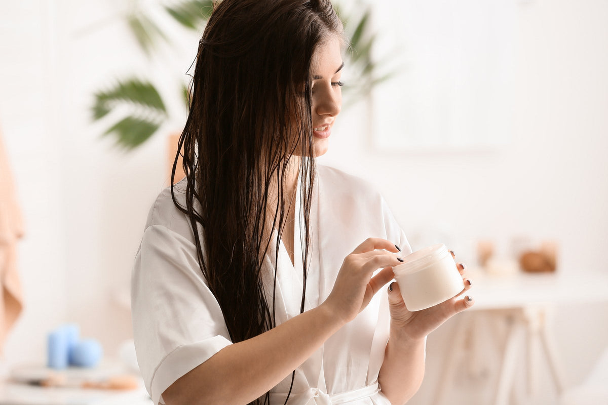 A woman in a white robe applying a hair mask from a jar to her long dark hair